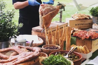 Un cuisinier découpe un jambon sur un buffet extérieur garni de pains, légumes, olives et charcuteries.