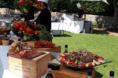 Buffet en plein air avec charcuterie, fruits, et bouteilles de vin, sous des parasols dans un jardin verdoyant.