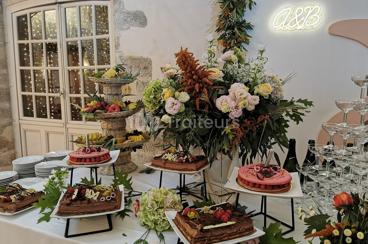 Table décorée avec des fleurs, des gâteaux variés et des fruits, entourée de verres et d'une lumière néon ’A&B’.