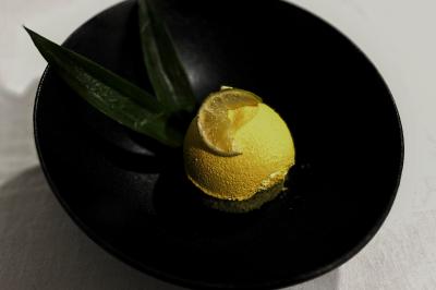 Dessert jaune en forme de dôme, garni d'une tranche de citron et accompagné de feuilles vertes, présenté dans une assiette…