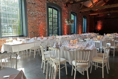 Traiteur France Evenement Traiteur Comines #4 Salle de réception avec tables rondes dressées, chaises blanches et murs en briques apparentes sous un toit en poutres.