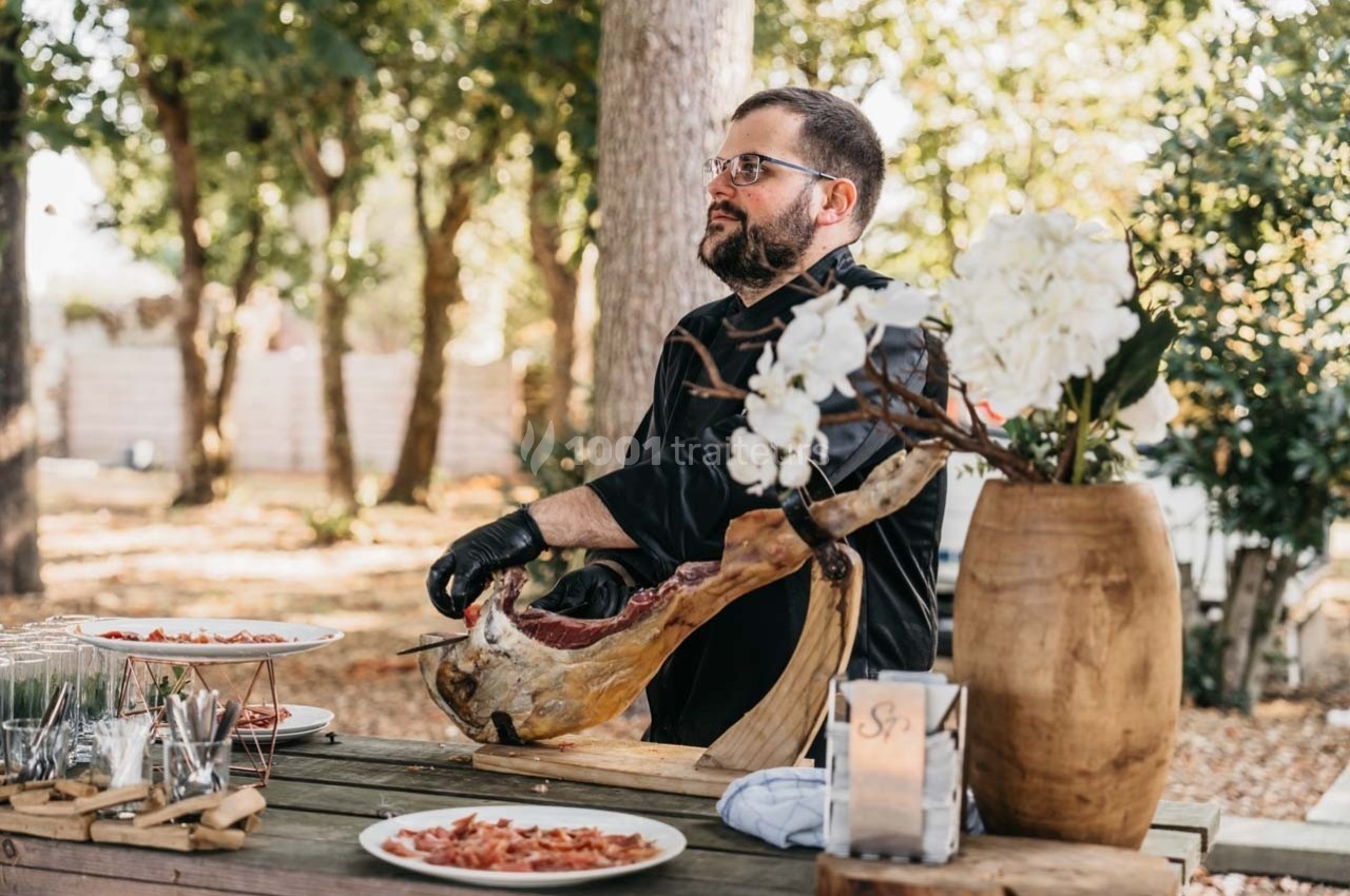 Un homme découpe un jambon cru sur une table en bois en extérieur, entouré d'arbres et de vaisselle.