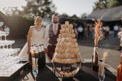 Tour de macarons sur une table de réception en extérieur, avec un couple en arrière-plan et des bouteilles de champagne.