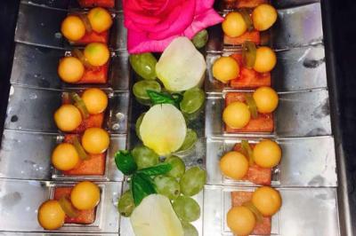 Traiteur Vironneau Evénementiel Galgon #5 Plateau décoratif avec des morceaux de melon, raisins, cubes de pastèque et une rose rose au centre.