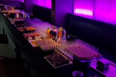 Buffet avec verrines, amuse-bouches, boissons et desserts disposés sur une longue table éclairée par une lumière violette.