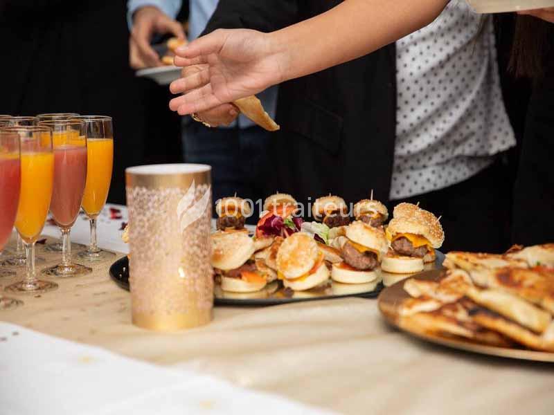 Plateau de mini-burgers et verres de cocktails sur une table lors d'un buffet, avec une main se servant.