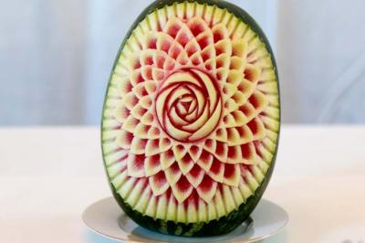 Pastèque sculptée avec des motifs floraux détaillés, posée sur une assiette blanche devant un fond neutre.