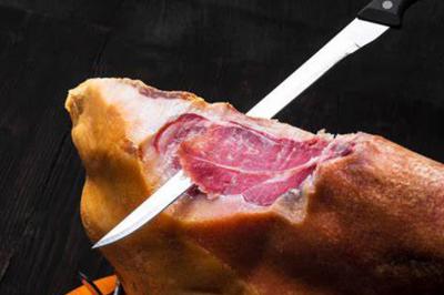 Un couteau tranche une pièce de jambon cru sur un fond sombre.