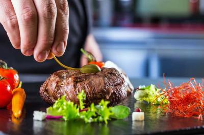 Traiteur Rubis Receptions Magny-le-Hongre #5 Une main dépose une sauce sur un steak accompagné de légumes et de garnitures colorées sur une assiette noire.