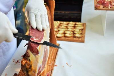 Une personne découpe un jambon sec sur un support en bois, avec des amuse-bouches disposés en arrière-plan.