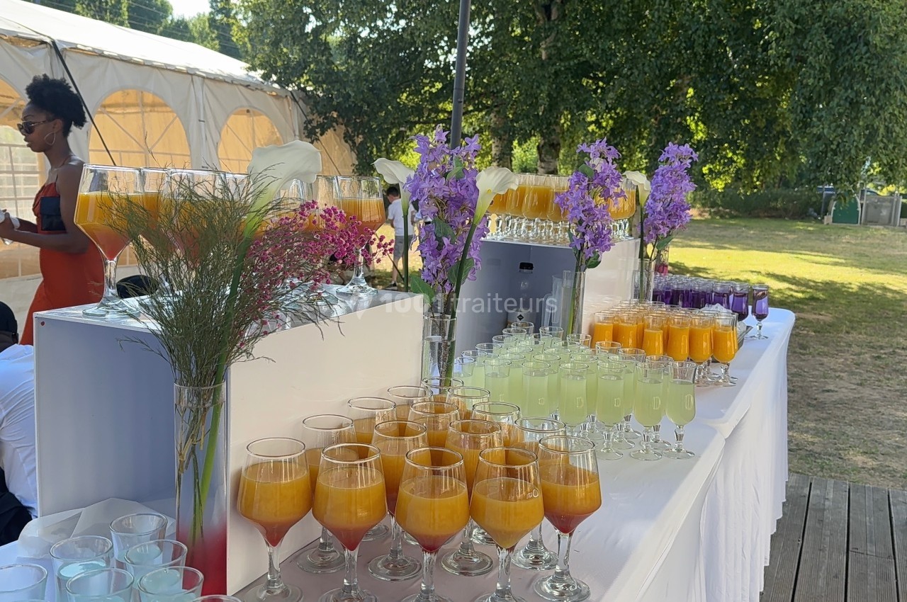 Table dressée en extérieur avec des verres de cocktails colorés, fleurs décoratives et tente en arrière-plan.