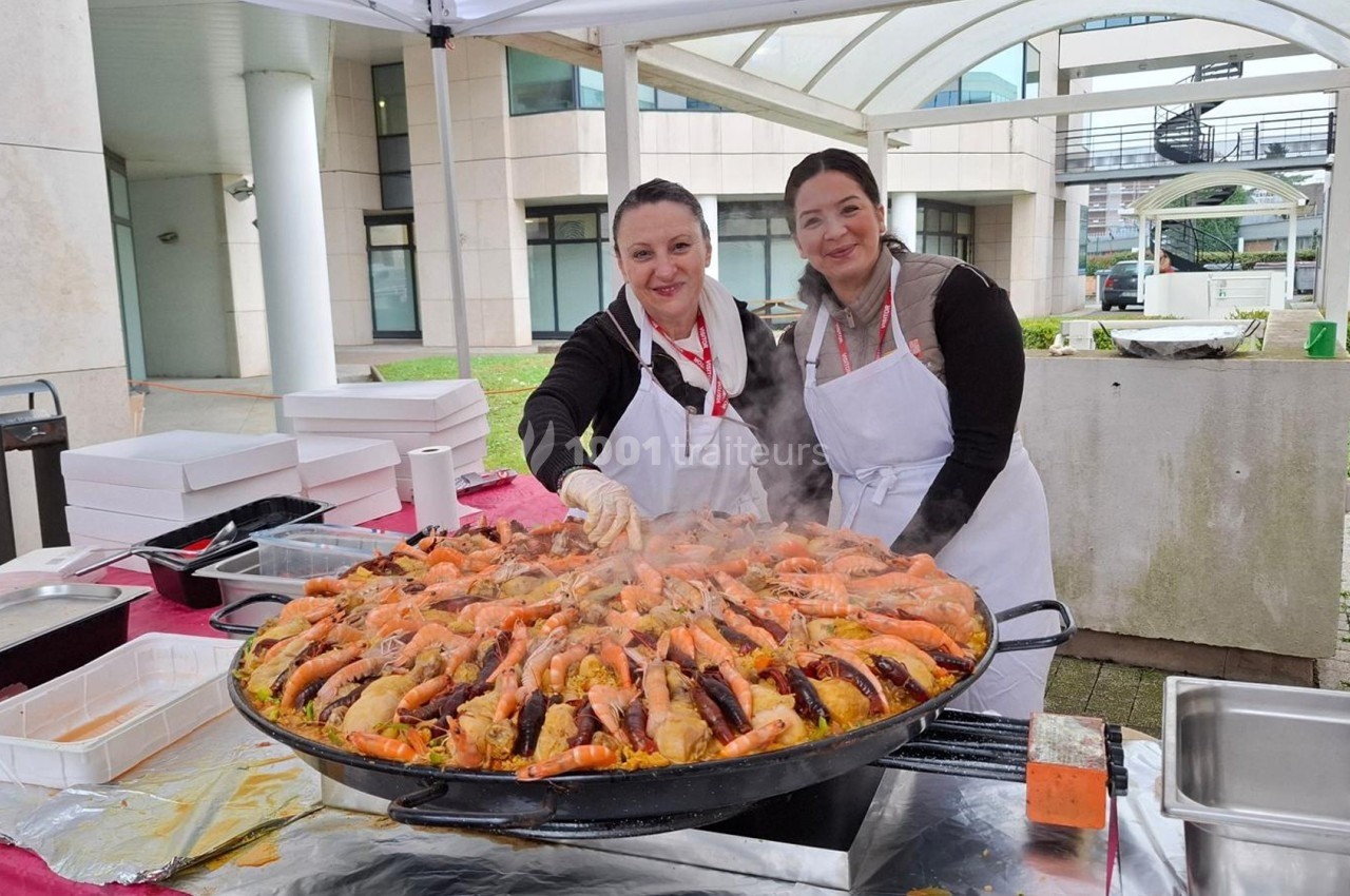 Traiteur Le Carré d'Or Traiteur Paris 15 #1 Deux femmes en tablier blanc cuisinent une grande paella fumante sous un chapiteau en extérieur.