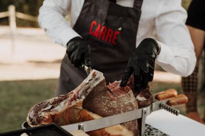 Un cuisinier en tablier découpe un jambon sec à l'aide d'un couteau sur un support en extérieur.