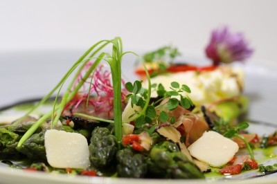 Assiette garnie d'asperges, copeaux de fromage, légumes colorés et herbes fraîches, avec un plat en arrière-plan.