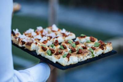 Plateau de focaccia garnie de morceaux de viande, herbes fraîches et fromage, tenu par une personne en chemise blanche.