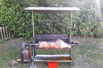 Un barbecue à rôtissoire en plein air avec des morceaux de viande en cuisson devant une haie verte.