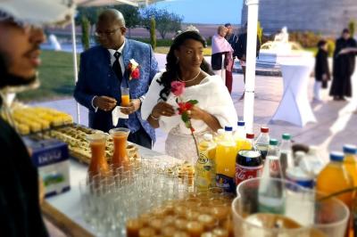 Un couple en tenue de mariage se sert à un buffet en plein air, entouré de boissons et de verrines.