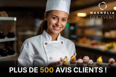 Une pâtissière souriante présente un plateau de petits gâteaux dans une cuisine professionnelle éclairée.