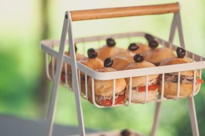 Plateau en métal contenant des mini-burgers garnis de tomates, olives noires et viande, posé en extérieur.