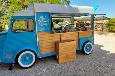 Camion bleu aménagé en food truck avec comptoir en bois, stationné sur un sol gravillonné près d'arbres.