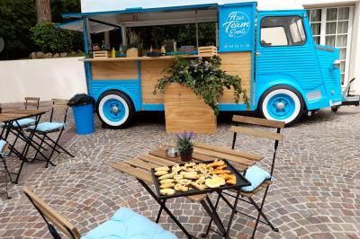 Camion bleu aménagé en food truck, stationné sur une place pavée avec tables et plateau de snacks en premier plan.