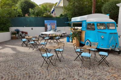 Camion bleu aménagé en food truck avec tables et chaises sur une terrasse pavée, voiture blanche en arrière-plan.