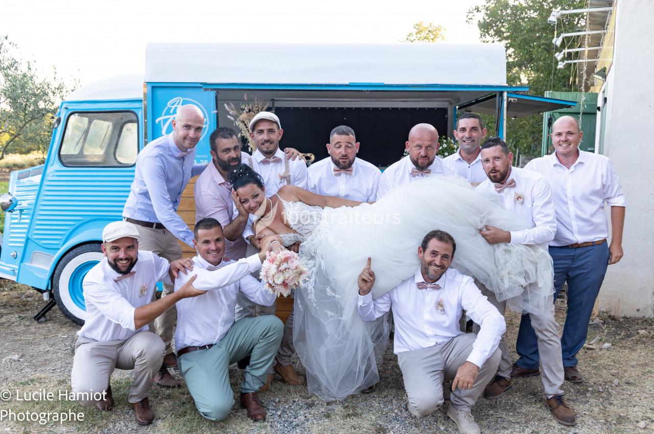 Un groupe d'hommes en chemises blanches porte une mariée souriante devant une camionnette bleue.