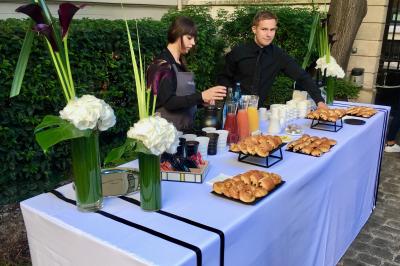 Table de buffet en extérieur avec viennoiseries, jus de fruits, café et fleurs, servie par deux personnes.