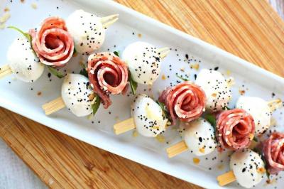 Brochettes apéritives avec mozzarella, tranches de charcuterie en forme de rose, graines et herbes sur un plateau.