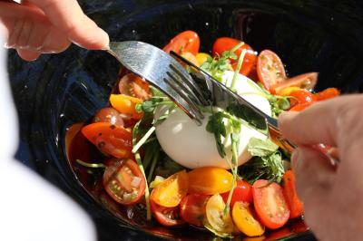 Traiteur Restaurant la Touloubre La Barben #3 Personne coupant une burrata garnie de tomates cerises colorées et de jeunes pousses dans une assiette noire.