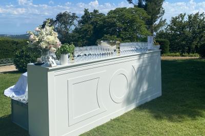 Bar extérieur blanc sur une pelouse, décoré de fleurs et de verres, entouré d'arbres sous un ciel ensoleillé.