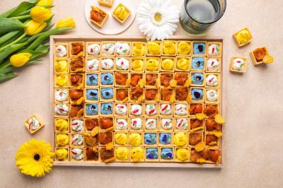Plateau de bouchées apéritives colorées disposées en rangées, entouré de fleurs jaunes et d'un verre d'eau.
