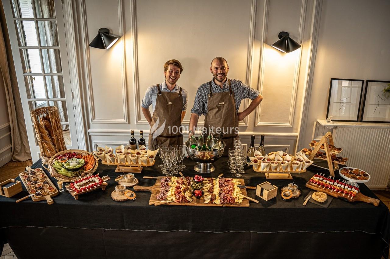 Deux hommes souriants en tabliers derrière une table garnie de plats variés, boissons et verrines dans une salle élégante.
