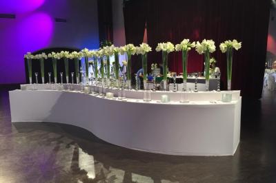 Traiteur Vip Traiteur Yerres #2 Alignement de vases transparents contenant des fleurs blanches sur une table blanche courbée dans une salle événementielle.