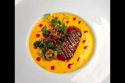 Plat gastronomique composé d'un morceau de viande grillée, sauce jaune, herbes fraîches et fleurs comestibles.
