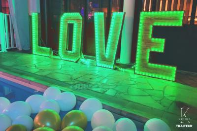 Lettres lumineuses formant le mot ’LOVE’ éclairées en vert, entourées de ballons blancs et dorés près d'une piscine.