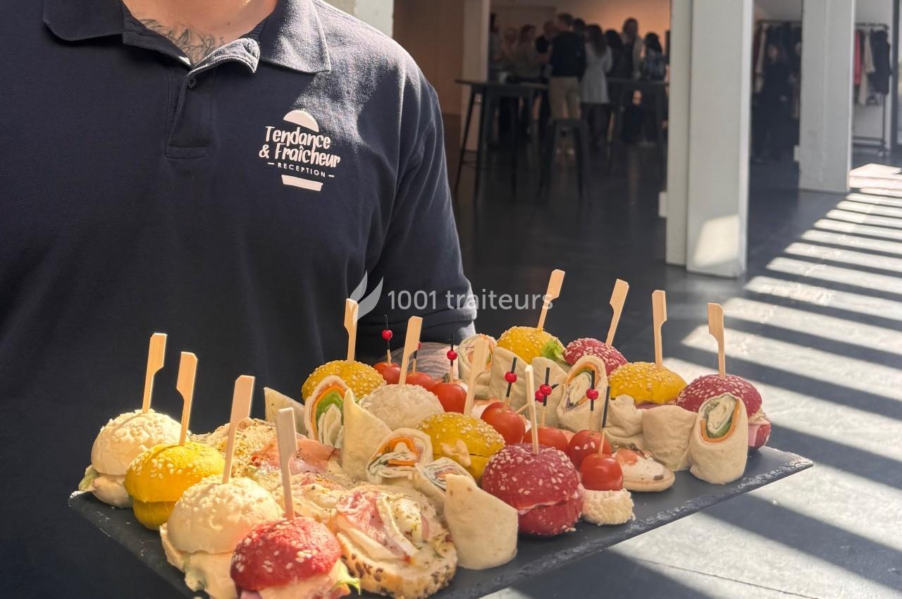 cocktail dinatoire Plateau de bouchées apéritives variées présentées par un serveur dans un espace lumineux avec des invités en arrière-plan.