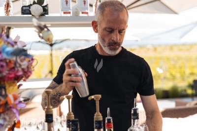 Bar à Cocktails Un barman prépare un cocktail en utilisant un shaker derrière un comptoir extérieur, entouré de bouteilles et d'ombrelles.