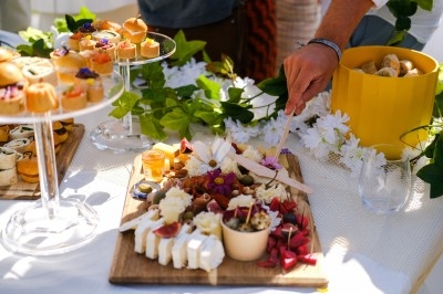 Buffets Cocktails Plateau de fromages et apéritifs variés sur une table décorée de fleurs et de verdure, avec une main servant un plat.