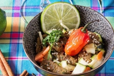 Bol de légumes frais avec citron vert, piment rouge, graines et herbes, sur une nappe colorée à carreaux.