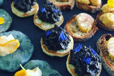 Assortiment de canapés garnis de tapenade, fleurs comestibles et autres ingrédients sur une table sombre.