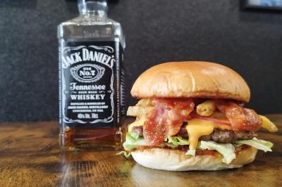 Burger garni de fromage, bacon et légumes, posé sur une table en bois, avec une bouteille de whisky en arrière-plan.