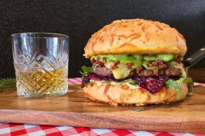 Burger garni de viande, fromage, guacamole et sauce, servi sur une planche avec un verre de whisky.