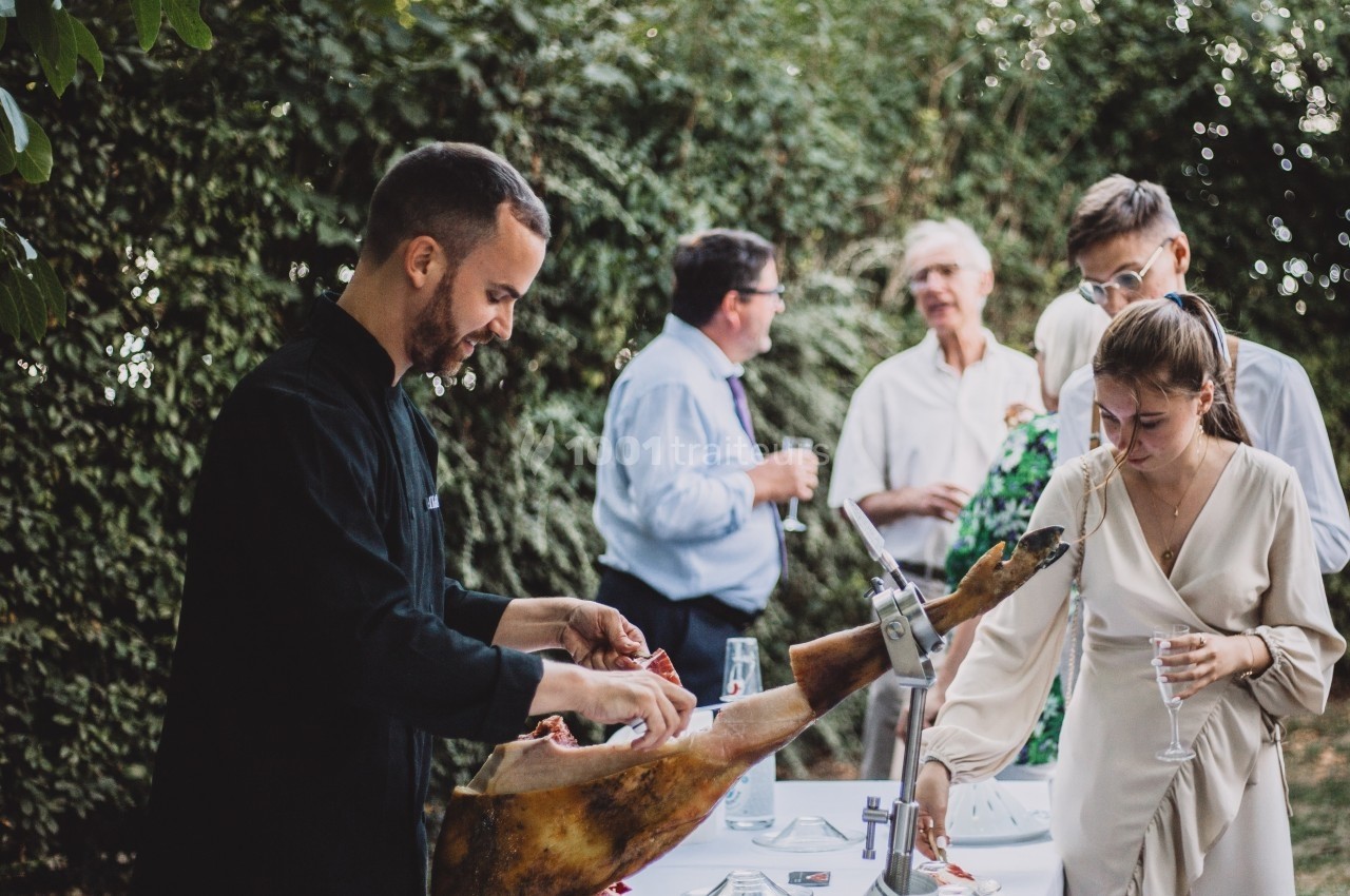 Un homme découpe du jambon sur un buffet en plein air lors d'un événement, entouré de convives discutant.