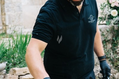 Traiteur Traiteur D'azur Saint-Vallier-de-Thiey #5 Un cuisinier en tenue noire prépare des légumes dans une poêle en extérieur, devant un mur en pierre et des fleurs.