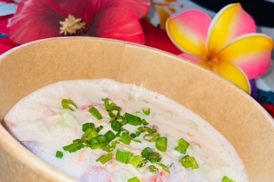 Salade tahitienne Bol en carton rempli d'une préparation crémeuse garnie de ciboulette, avec des fleurs tropicales en arrière-plan.