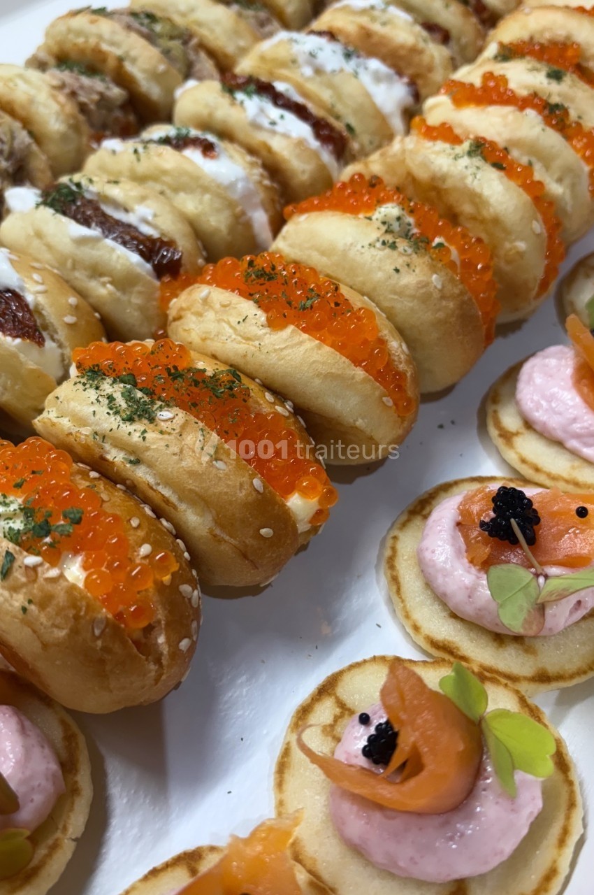 Assortiment de mini-sandwichs garnis d'œufs de poisson et de blinis au saumon et crème rose, disposés sur un plateau.