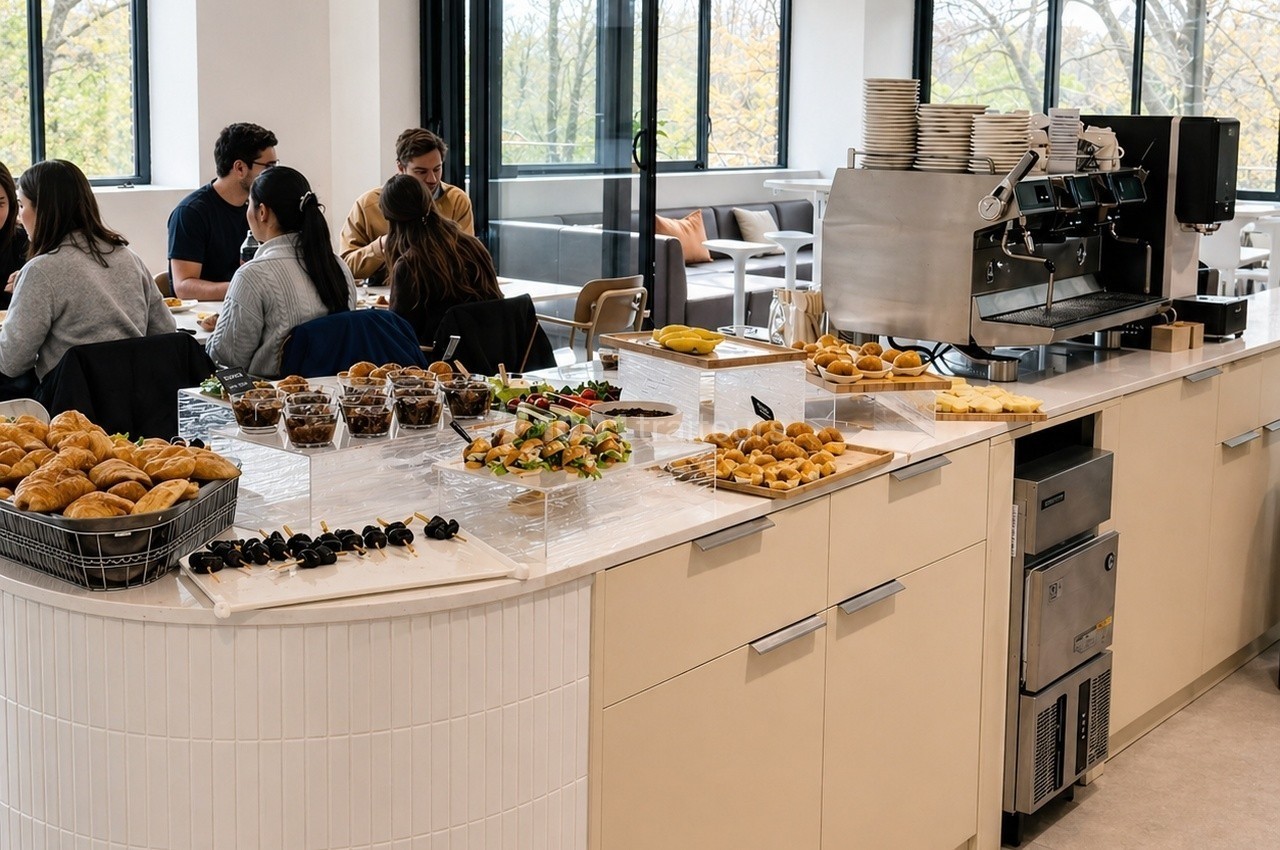 Buffet avec viennoiseries, fruits, brochettes et machine à café dans un espace lumineux avec des convives assis.