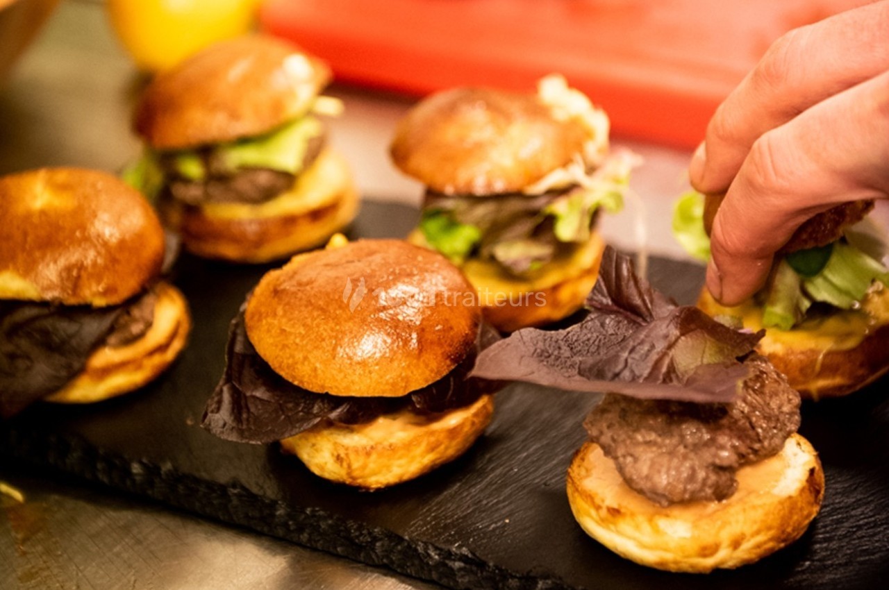 Traiteur La Tricoterie Traiteur Saint-Gilles #1 Mini-burgers garnis de viande, salade et sauce, disposés sur une ardoise, avec une main ajustant la garniture.