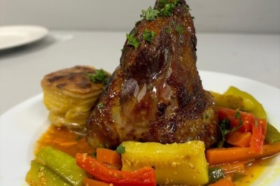 Jarret d'agneau braisé servi avec des légumes colorés et un gratin de pommes de terre, présenté sur une assiette blanche.